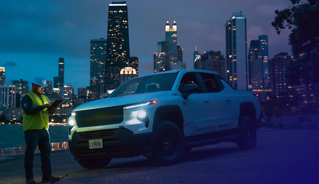 an utility worker in a hard hat and high visibility vest reviews a tablet beside a branded electric pickup truck at night with the chicago skyline illuminated in the background