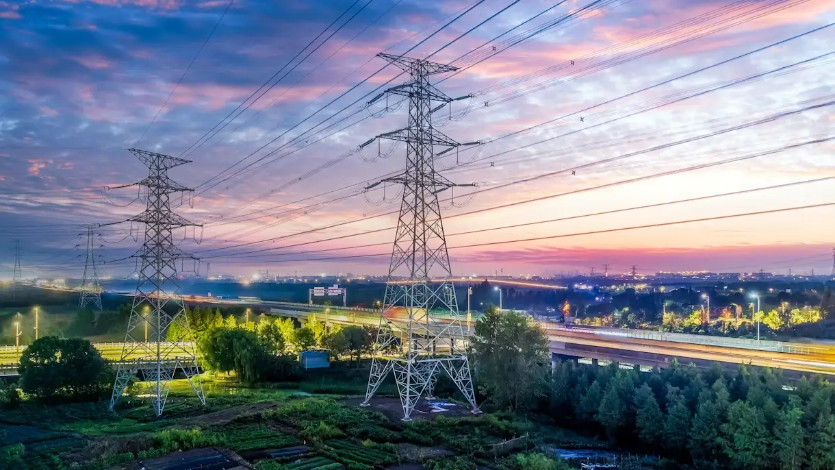 aerial view of high voltage electricity transmission towers and power lines at dusk with a colorful sky and illuminated highway infrastructure in the background