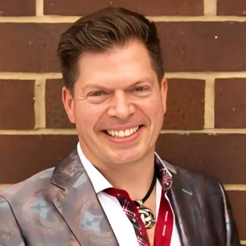 smiling man in a patterned blazer and white shirt posing against a brick wall looking at the camera