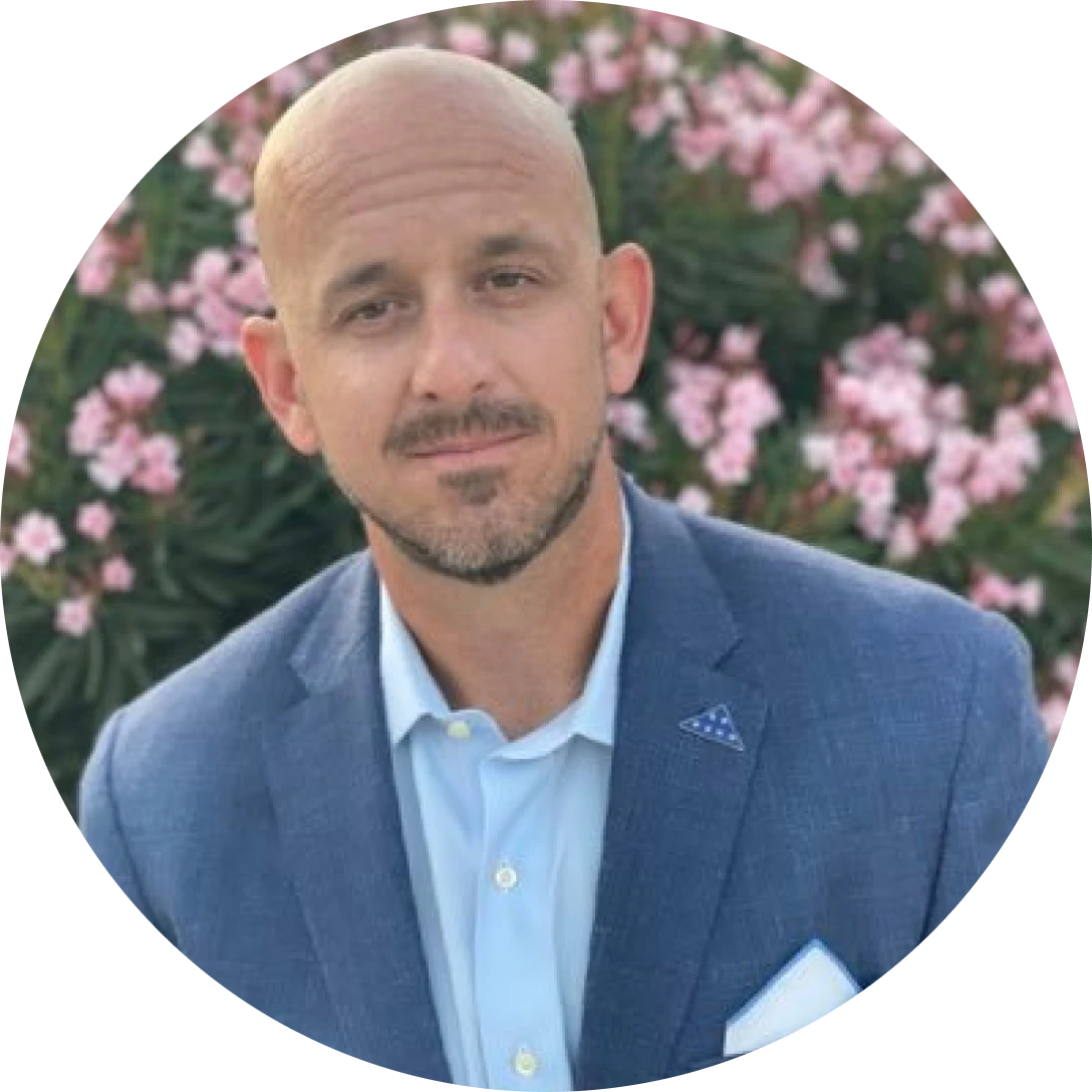 portrait of a bald man with a short beard in a blue blazer and light blue shirt against pink flowering bushes behind him